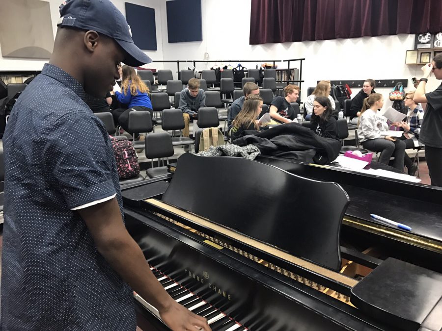 Shows a student auditioning for the musical playing the piano to learn the right pitch for the music they are given, and other students practicing their parts in the background, and interacting with each other.
