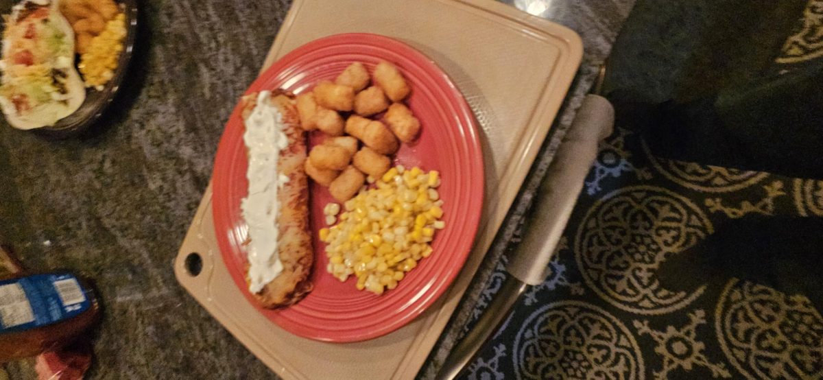 A plated dinner featuring baked chicken, potatoes, and corn is shown from above. The meal reflects a simple, homemade approach to cooking.