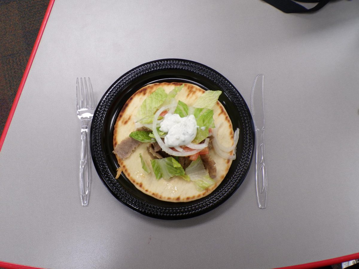 A gyro served on a plate, framed by silverware on either side. A straightforward presentation of lamb, pita, and toppings ready to eat.