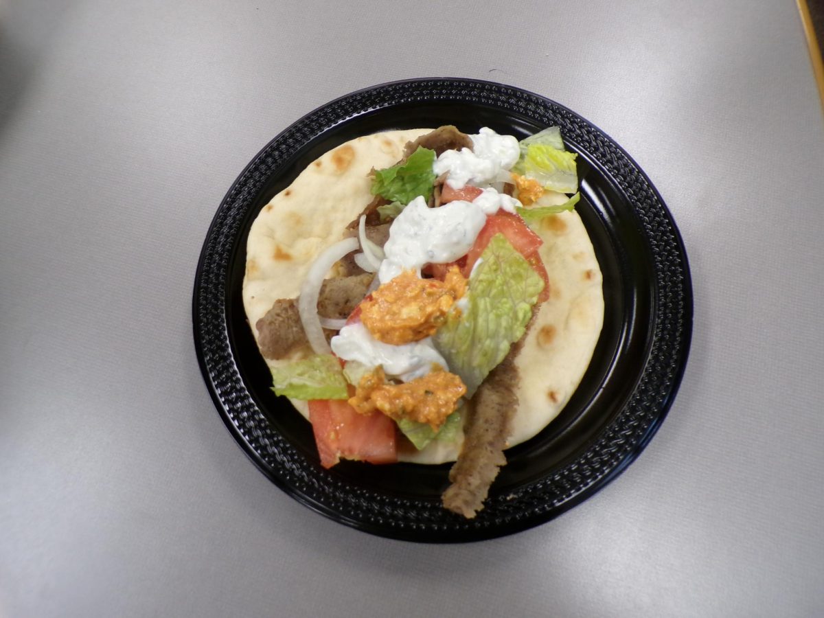 A close-up view shows a gyro fresh off the grill, with crisped meat and soft pita bread. The meal reflects traditional Greek street food commonly served in local restaurants.