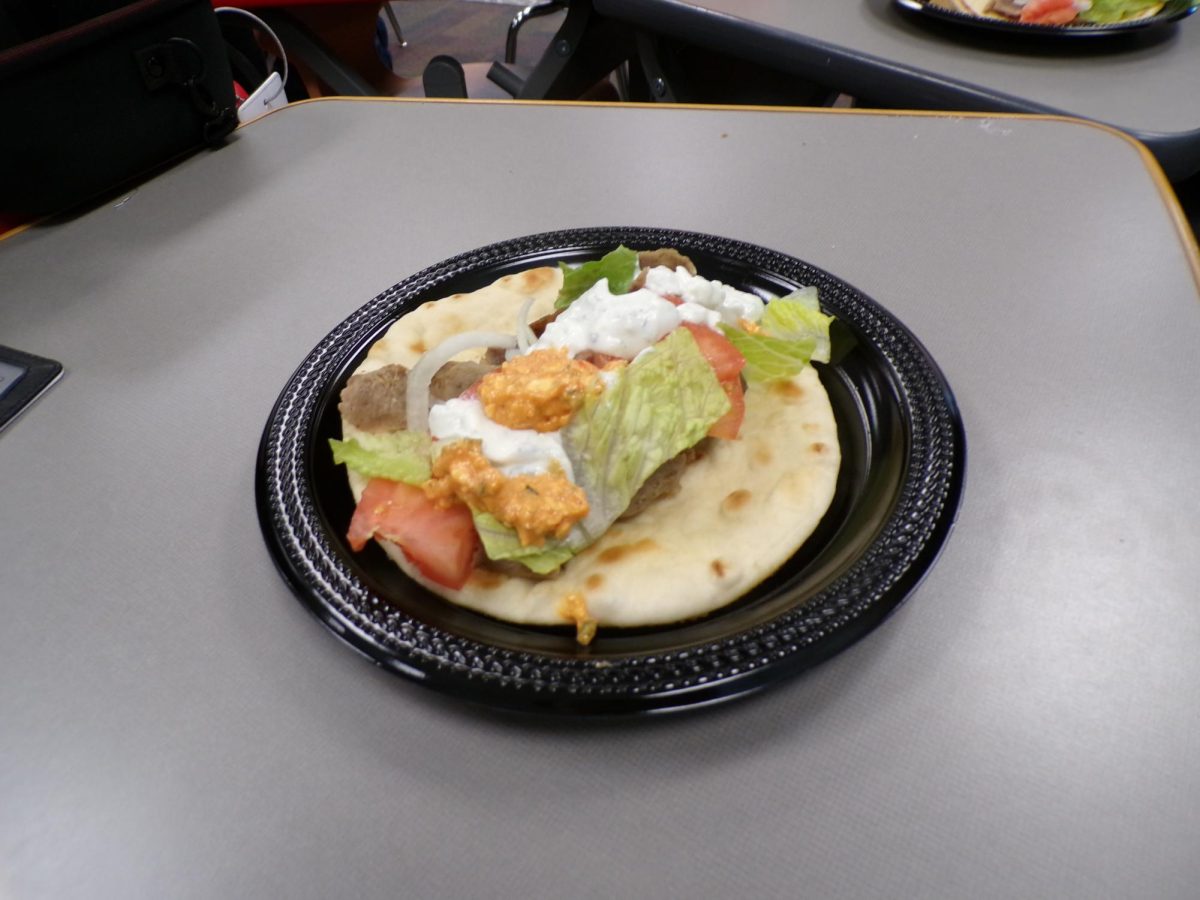 A gyro is prepared on a white paper wrap, layered with meat, fresh vegetables and sauce. The classic Greek dish combines warm and cool ingredients for balanced flavor.
