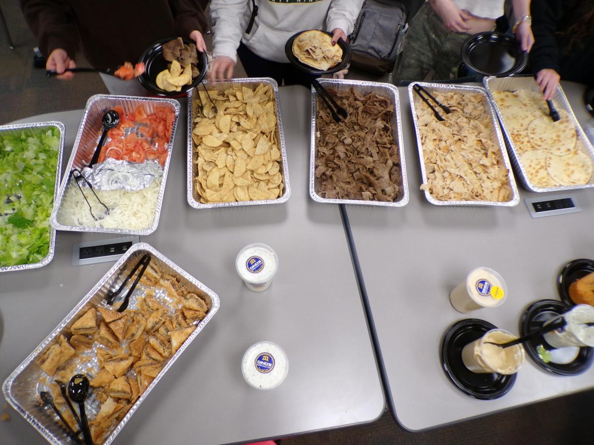 In this overhead view of the Mediterranean catering spread, students are pictured selecting from aluminum trays to build their own gyros. The table features a vibrant selection of fresh ingredients including lettuce, tomatoes, onions, pita bread, and gyro meat, alongside a large tray of golden-brown baklava.