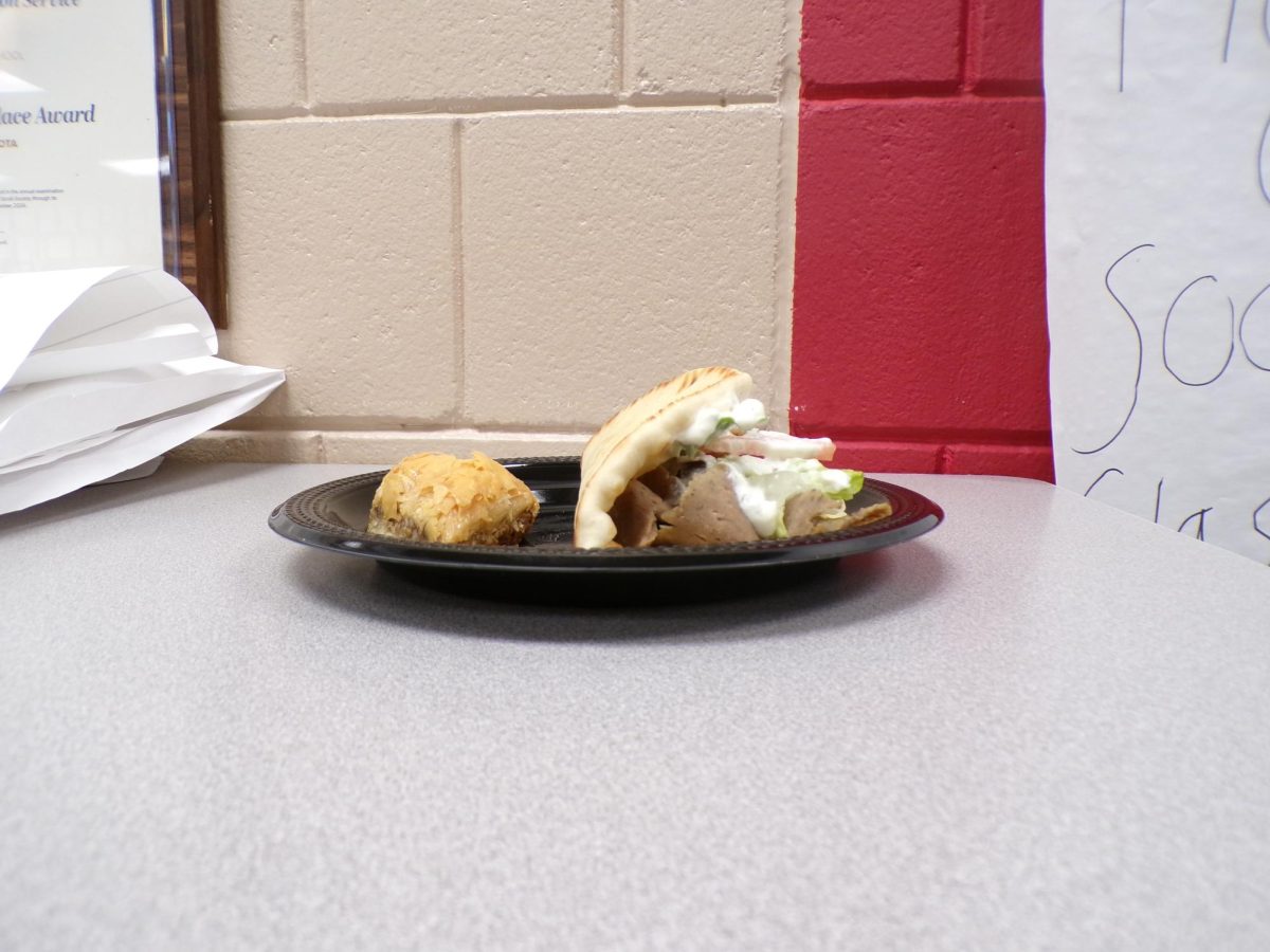A lamb gyro sits beside a small piece of baklava, neatly arranged on the table. The savory meat and tangy cucumber cucumber sauce contrast with the sticky sweetness and cinnamon-rich aroma of the dessert.
