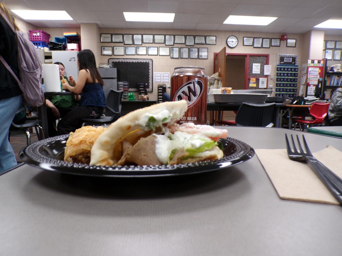 A close-up focuses on the gyro and baklava, with a can of root beer visible in the background. The creamy sauce and lamb pair with flaky, honeyed layers of baklava as the soda adds a cool, fizzy aftertaste.