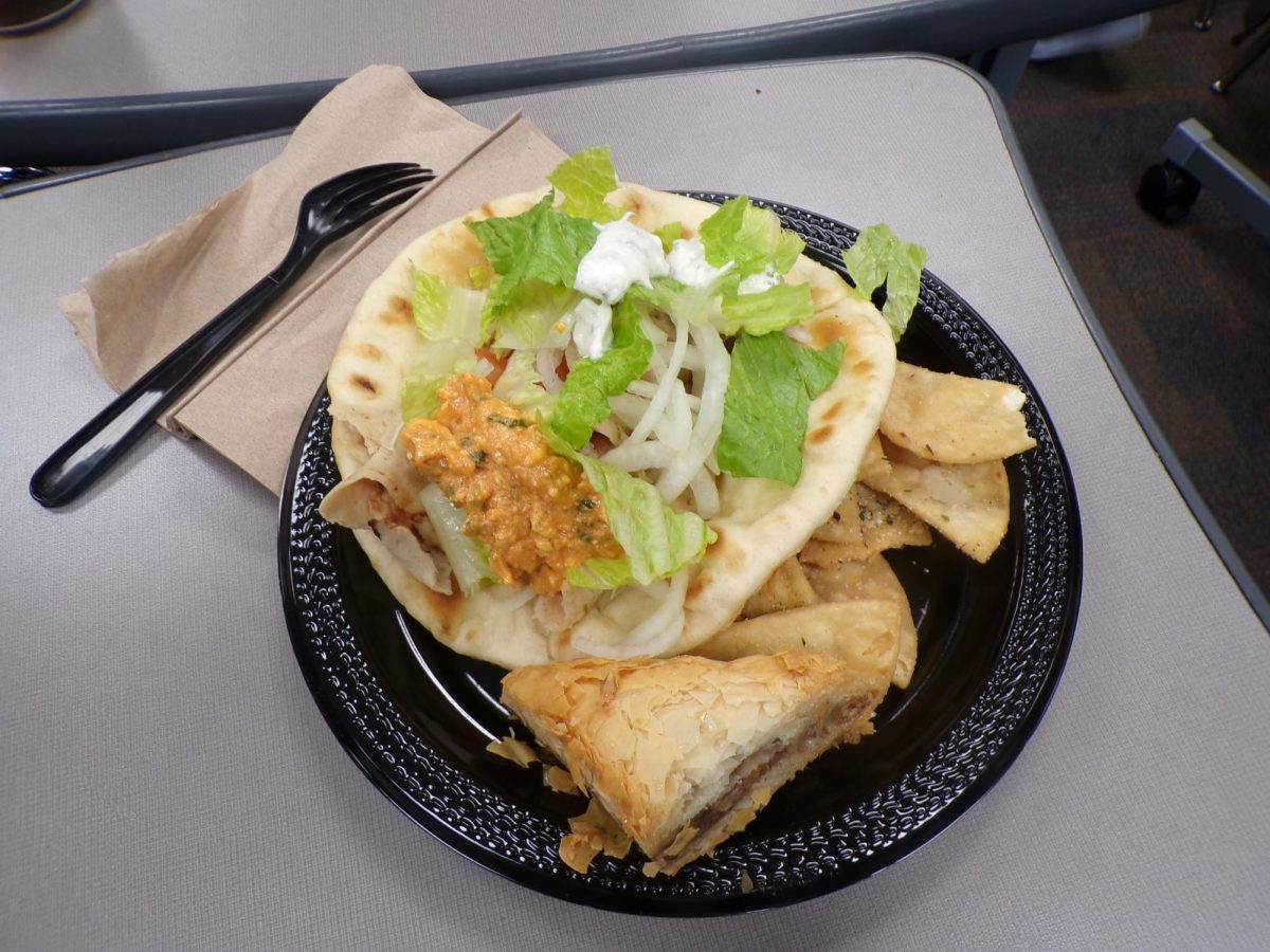 A vibrant, overstuffed gyro sits on the plate, wrapped in pita and layered with creamy sauces and crisp vegetables. With golden seasoned chips and a flaky, honey‑soaked baklava.