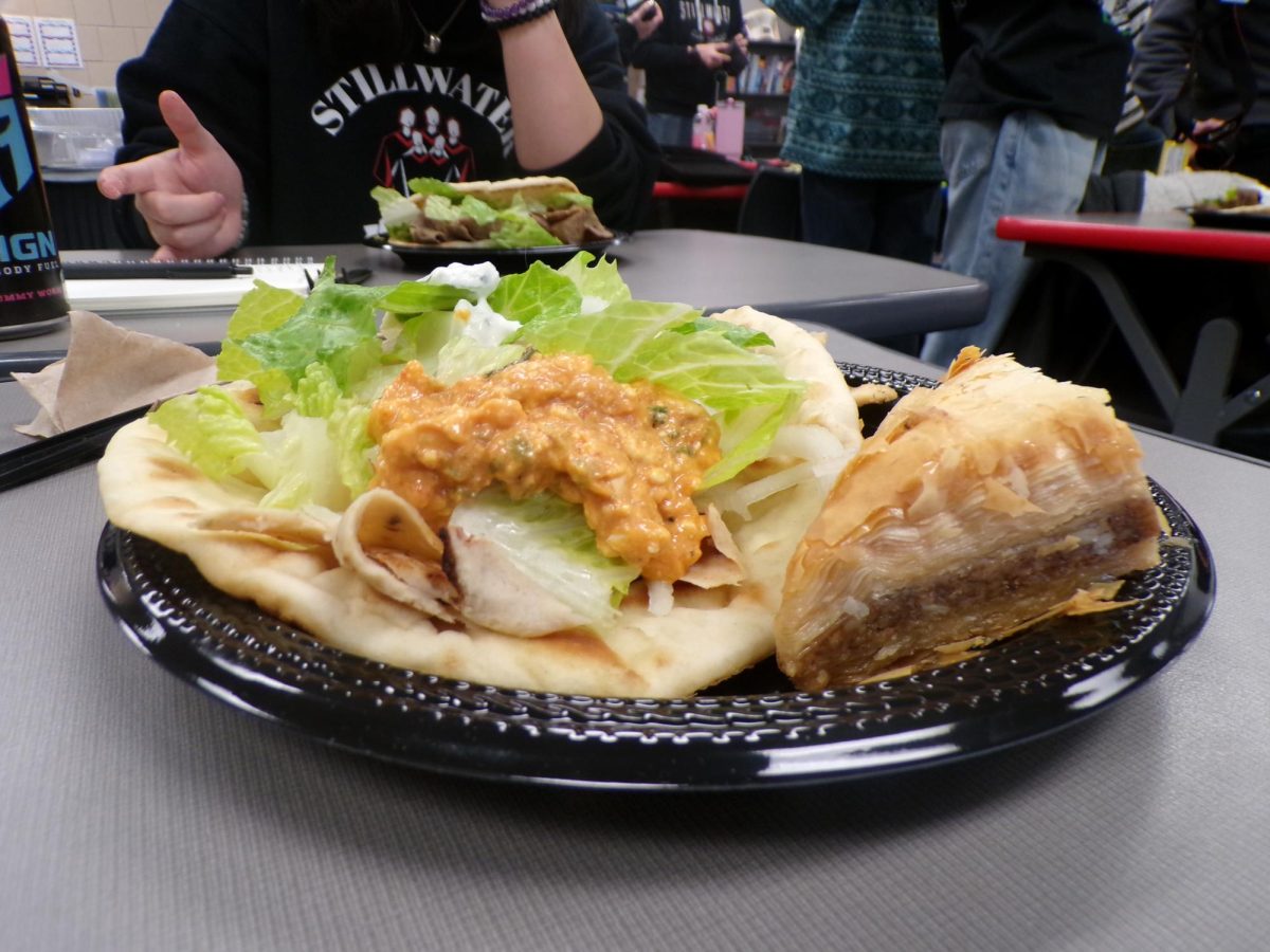 A warm chicken gyro sits on the plate with fresh lettuce and bold sauce. A piece of sweet, nutty baklava rests beside it.